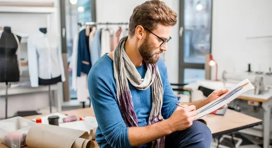 Un diseñador de producto con gafas y bufanda revisa bocetos de moda en su taller, rodeado de maniquíes y telas.