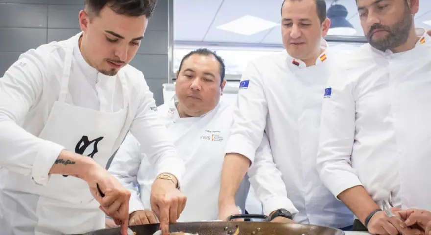 Cuatro chefs observan atentamente una paella. Se debate la diferencia entre chef y cocinero mientras preparan el plato.