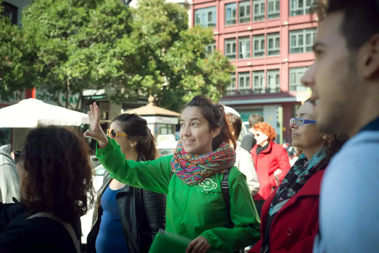 Joven guía sonriente, con bufanda colorida y chaqueta verde, saluda a un grupo. Podría ser una estudiante de grado superior de turismo en una visita guiada.