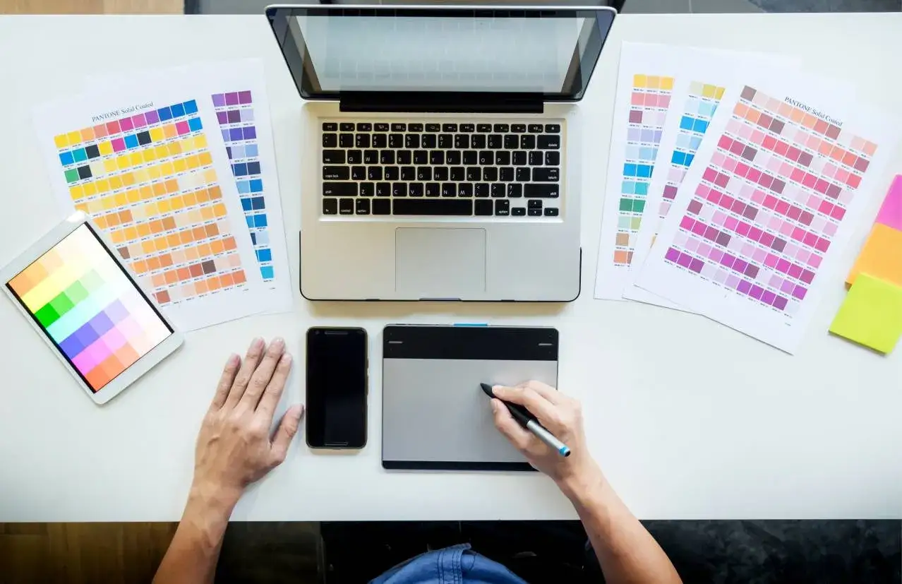 Dise&ntilde;ador gr&aacute;fico trabajando en su escritorio con paletas de colores, tablet, m&oacute;vil y port&aacute;til. As&iacute; se ve el d&iacute;a a d&iacute;a de cuanto gana un dise&ntilde;ador gr&aacute;fico.