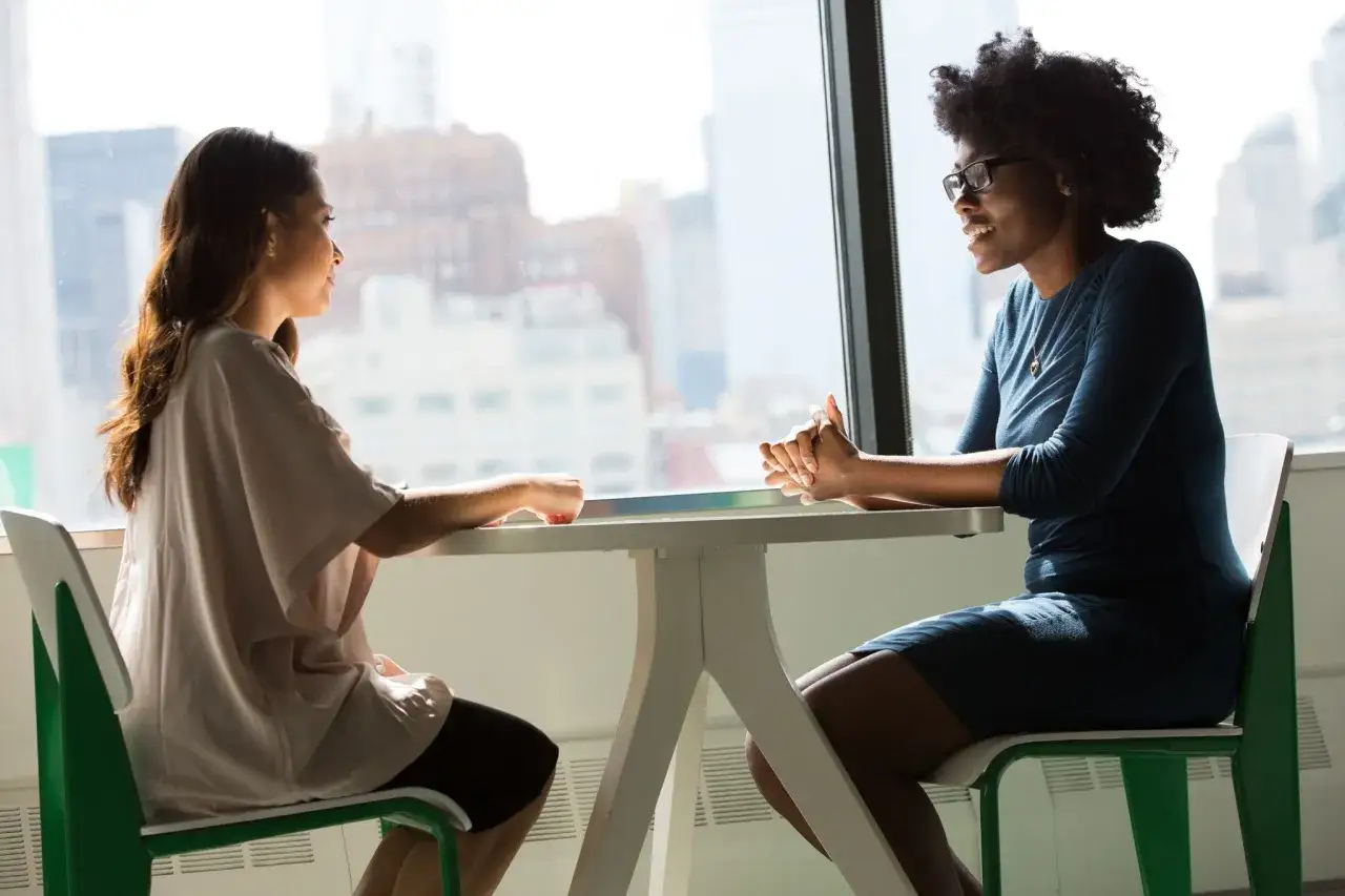 Dos mujeres conversan en una mesa, creando una imagen personal de conexi&oacute;n y di&aacute;logo.
