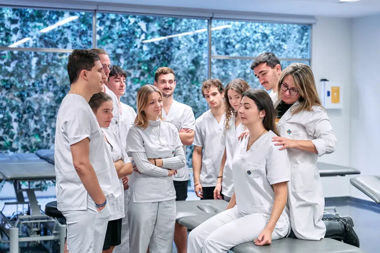 Estudiantes de fisioterapia aprenden técnicas de palpación en un aula. Un grupo diverso de jóvenes, con batas blancas, observa atentamente.