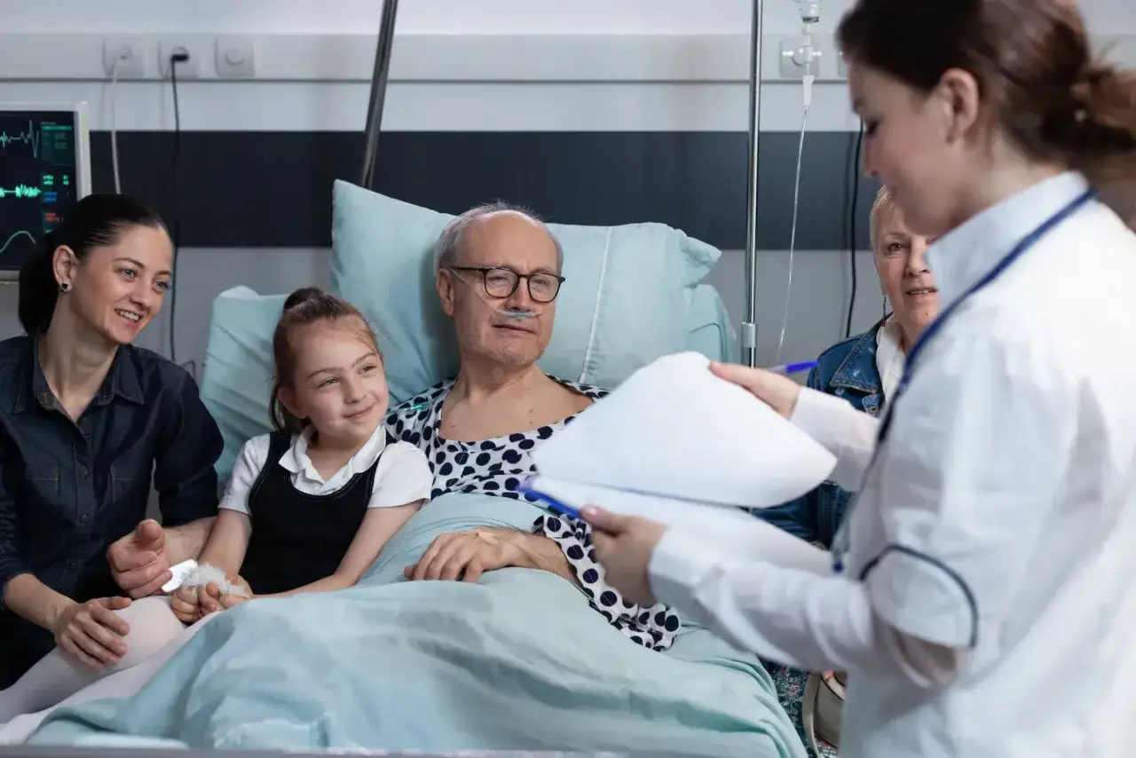 Paciente en cama de hospital con familia. Una auxiliar de enfermer&iacute;a, con su grado medio de auxiliar de enfermer&iacute;a, revisa un informe.