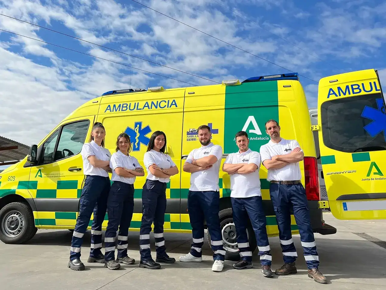 Equipo de fp técnico emergencias sanitarias posa orgulloso junto a una ambulancia amarilla y verde bajo un cielo azul con nubes.