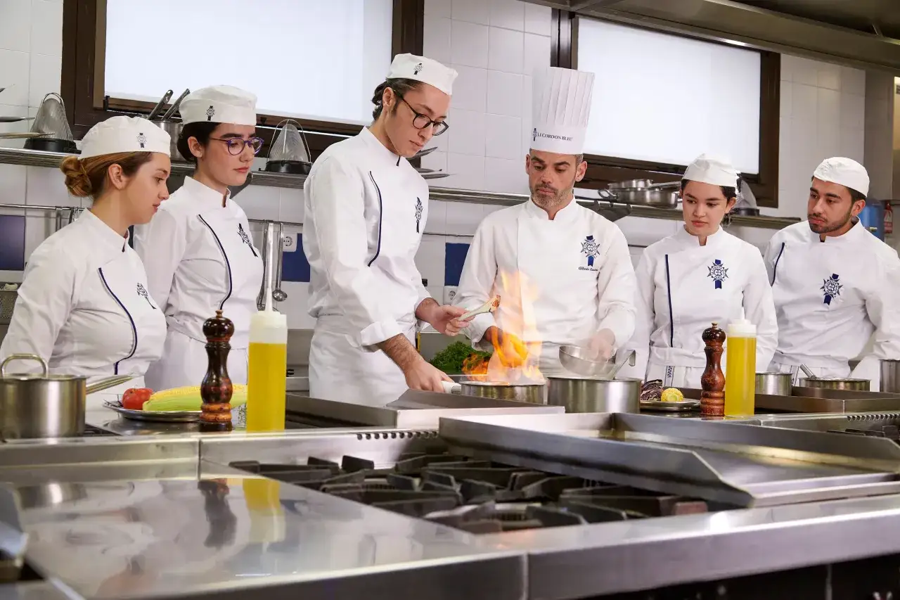 Estudiantes de carrera de gastronom&iacute;a aprenden t&eacute;cnicas culinarias en una cocina profesional, con un chef demostrando flambeado.