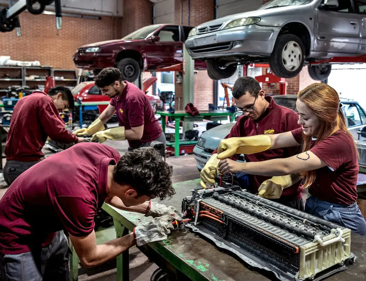 Estudiantes de grados medios en Barcelona trabajan en un taller mecánico, reparando un componente de coche.