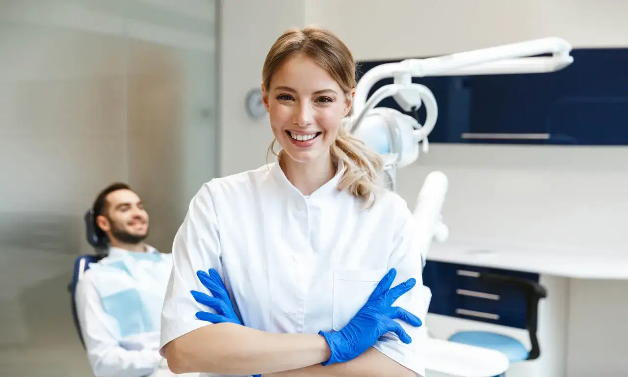 Sonrisa de profesional dental con guantes azules, lista para un curso de higiene bucodental. Paciente sonriente en silla dental al fondo.