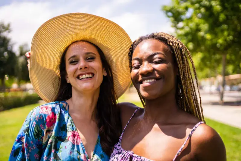 Dos amigas sonriendo bajo el sol, una con sombrero y vestido floral, la otra con trenzas. Una imagen personal de alegría compartida.
